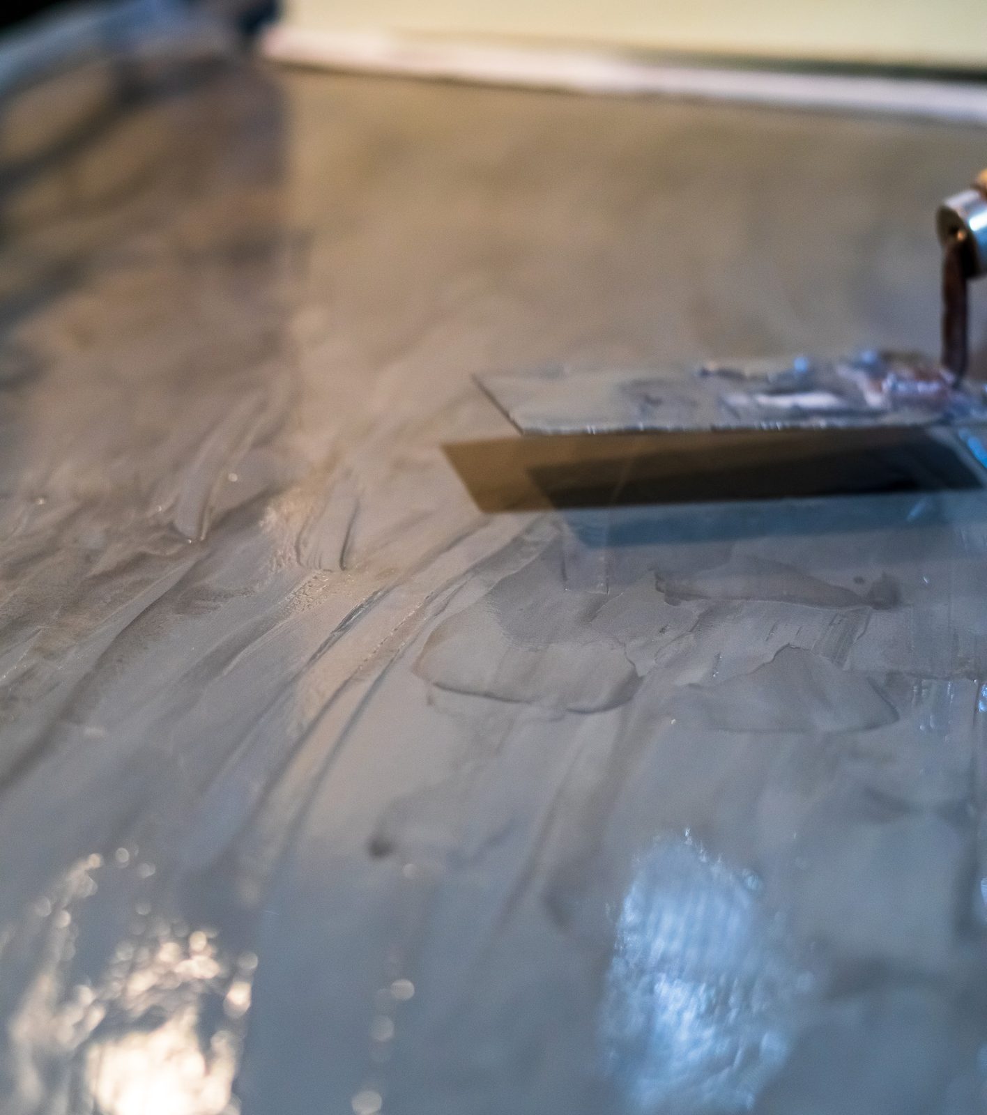Squeegee spreading a light floor coating over gray concrete in a sunlit room
