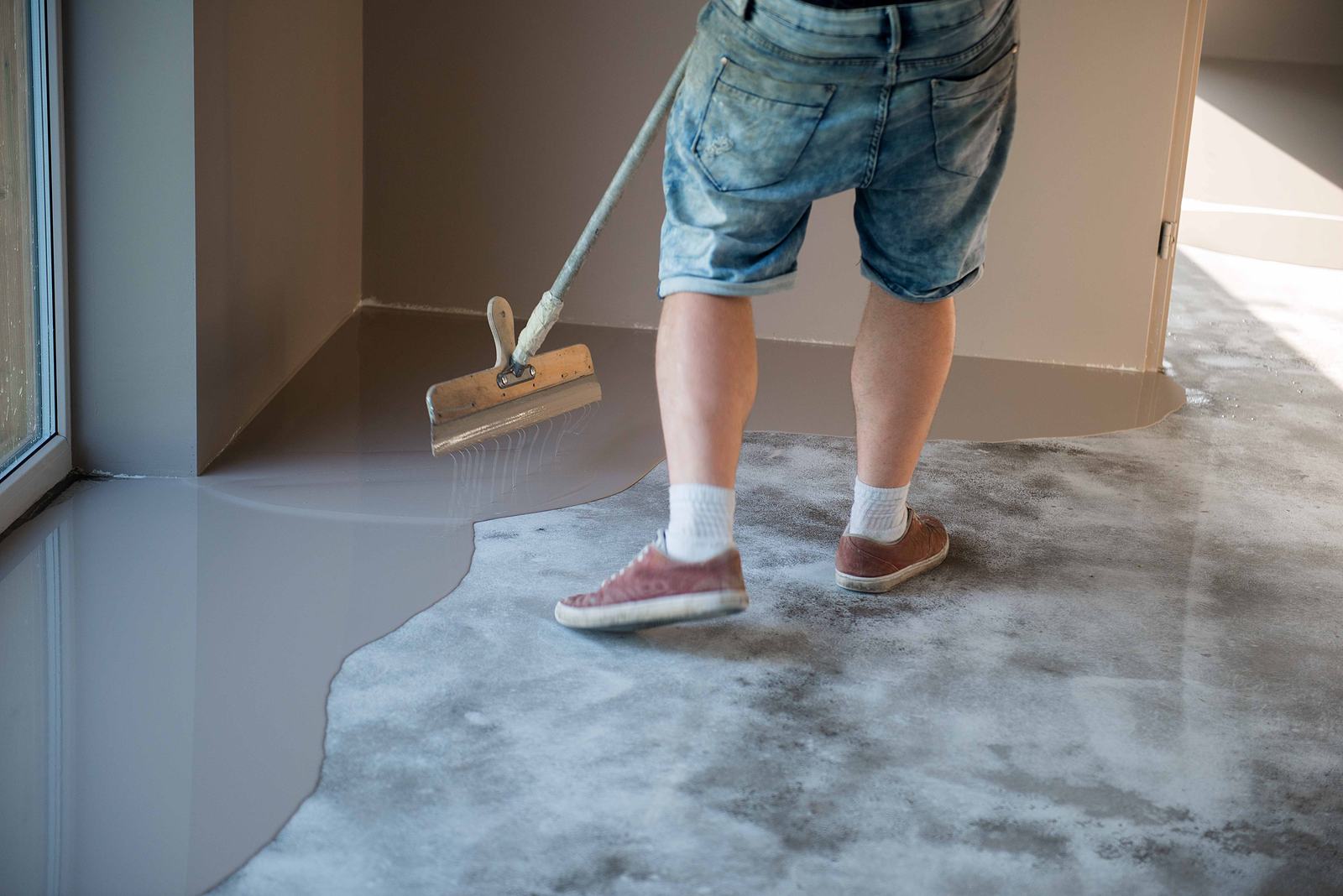 Worker spreading gray epoxy across an indoor concrete floor in an empty room